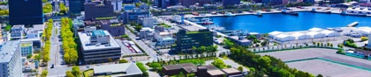 Cityscape of Chiba, Japan, featuring urban buildings, a waterfront harbor, and green spaces under a clear blue sky