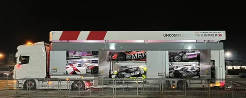 Scan Global Logistics motorsport transporter carrying multiple race cars in illuminated compartments at night.